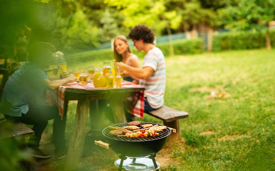 Een gezin eet samen in de tuin terwijl op de voorgrond een kleine barbecue ligt vol gegrilde spiesjes, maïs en hamburgers.