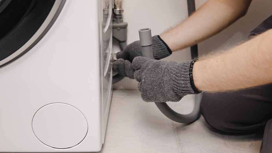 Een persoon met grijze handschoenen sluit een grijze slang aan op een witte wasmachine.
