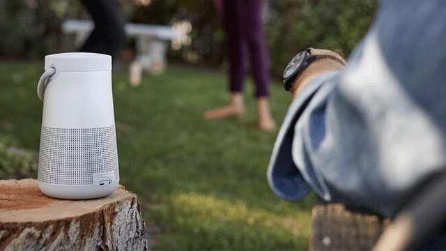 Witte speaker op een boomstronk, gazon op de achtergrond met vage figuren. Persoon met horloge zit ook op een boomstronk.