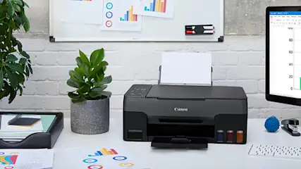 Een printer op een bureau met een whiteboard en grafieken aan de muur, naast een computer, planten en kantoorbenodigdheden.