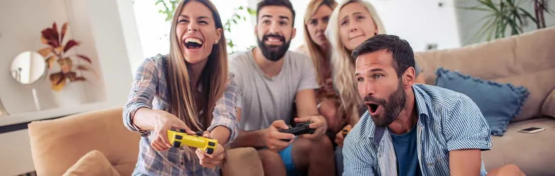 A group of friends is gathered on a sofa, enthusiastically enjoying a video game, all laughing and focused on the action.