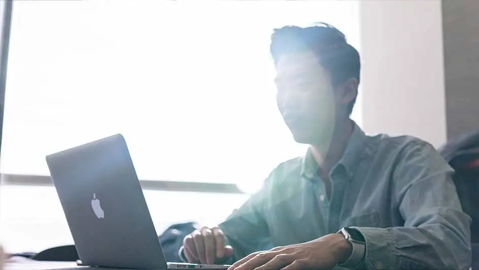 Person at desk using laptop. Open laptop with logo facing viewer. Person wears light shirt and watch. Window is in background.