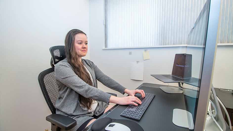 Een vrouw zit aan een bureau met een toetsenbord, muis en grote monitor in een kantooromgeving.