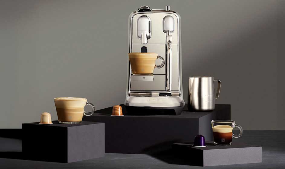 A stylish coffee machine stands on black blocks, with espresso cups, capsules and a metal milk jug against a grey background.