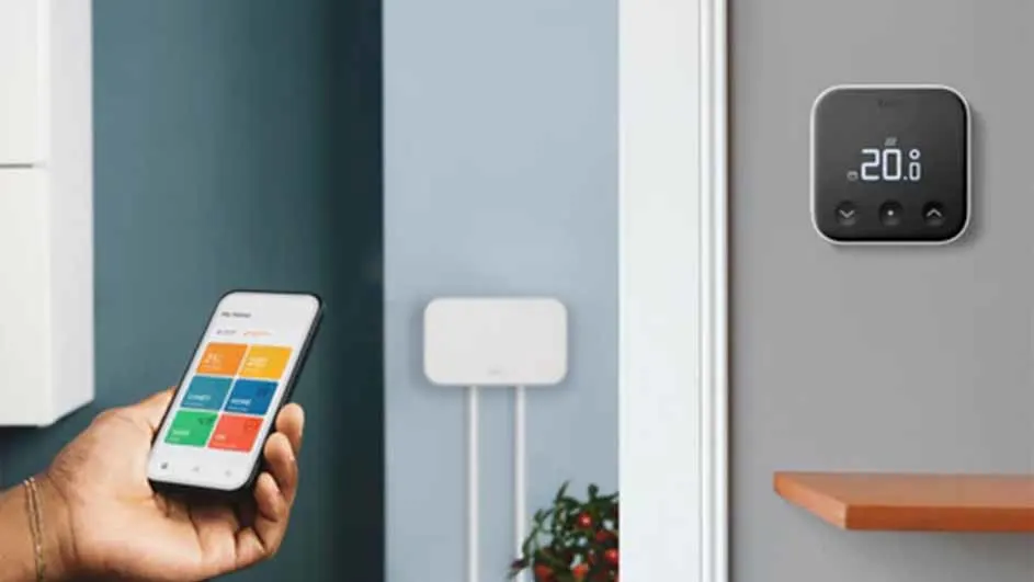 Hand houdt smartphone met app naast thermostaat aan grijze muur met houten plank. Witte unit met snoeren zichtbaar.