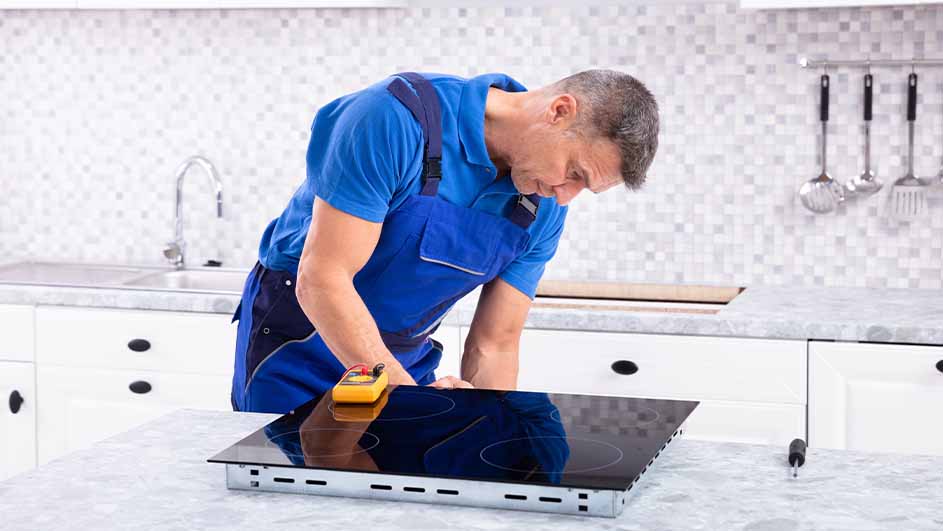 Een man in blauwe werkkleding inspecteert een zwarte kookplaat met een geel meetinstrument in een keuken.