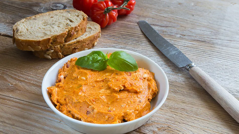 Op een houten tafel staan brood, tomaten, een mes en een witte kom met oranje smeersel, gegarneerd met groene basilicum.