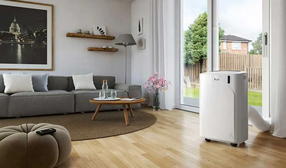 A white Pluïe portable air conditioner stands by a large window in a modern living room with a grey sofa, table, and soft wooden flooring.