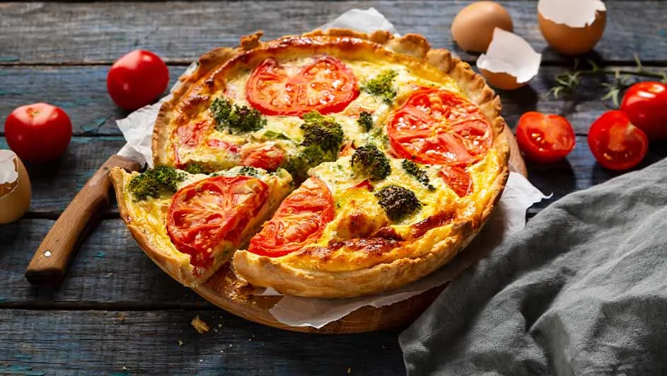 Een quiche met broccoli en tomaat, op een houten plank, met tomaten en eieren op een blauwe houten tafel.