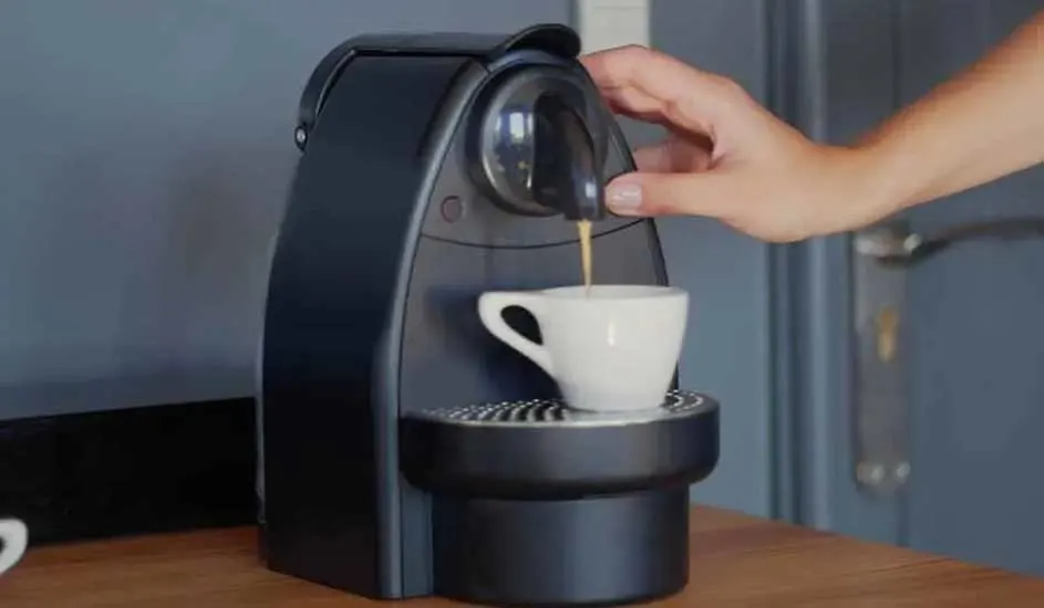 Een hand bedient een zwarte koffiecupmachine op een houten tafel, terwijl er koffie in een wit kopje stroomt tegen een grijze achtergrond.