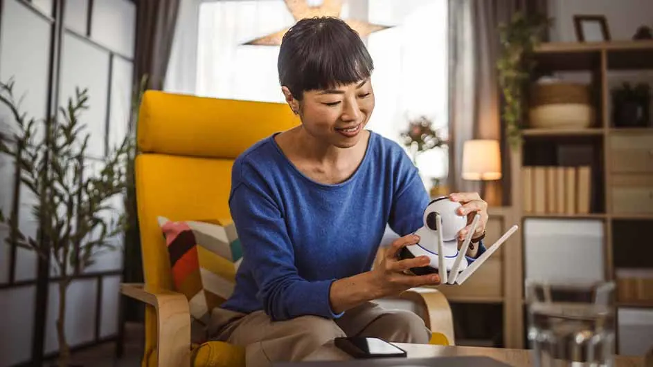 Vrouw zit in een gele stoel en bekijkt een witte beveiligingscamera. Op de tafel ligt een zwarte telefoon. Achtergrond: kast en gordijnen.
