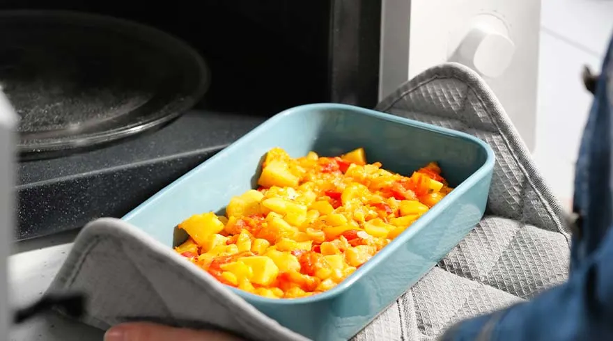 Een blauwe schaal met geel en rood voedsel in een magnetron, vastgehouden met een grijze ovenwant.