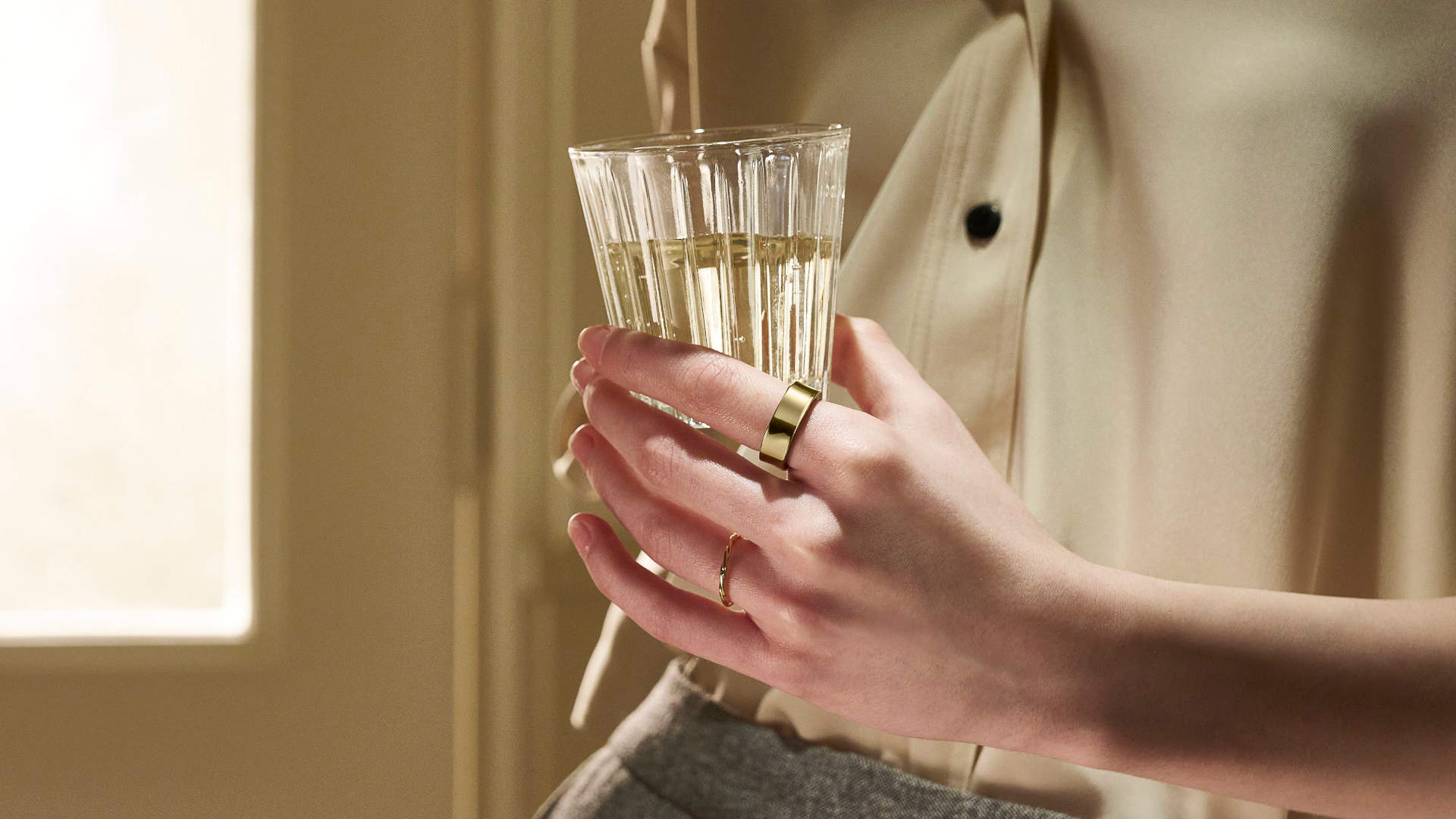 Een persoon houdt een glas met een heldere vloeistof vast, met gouden ringen om de vingers.