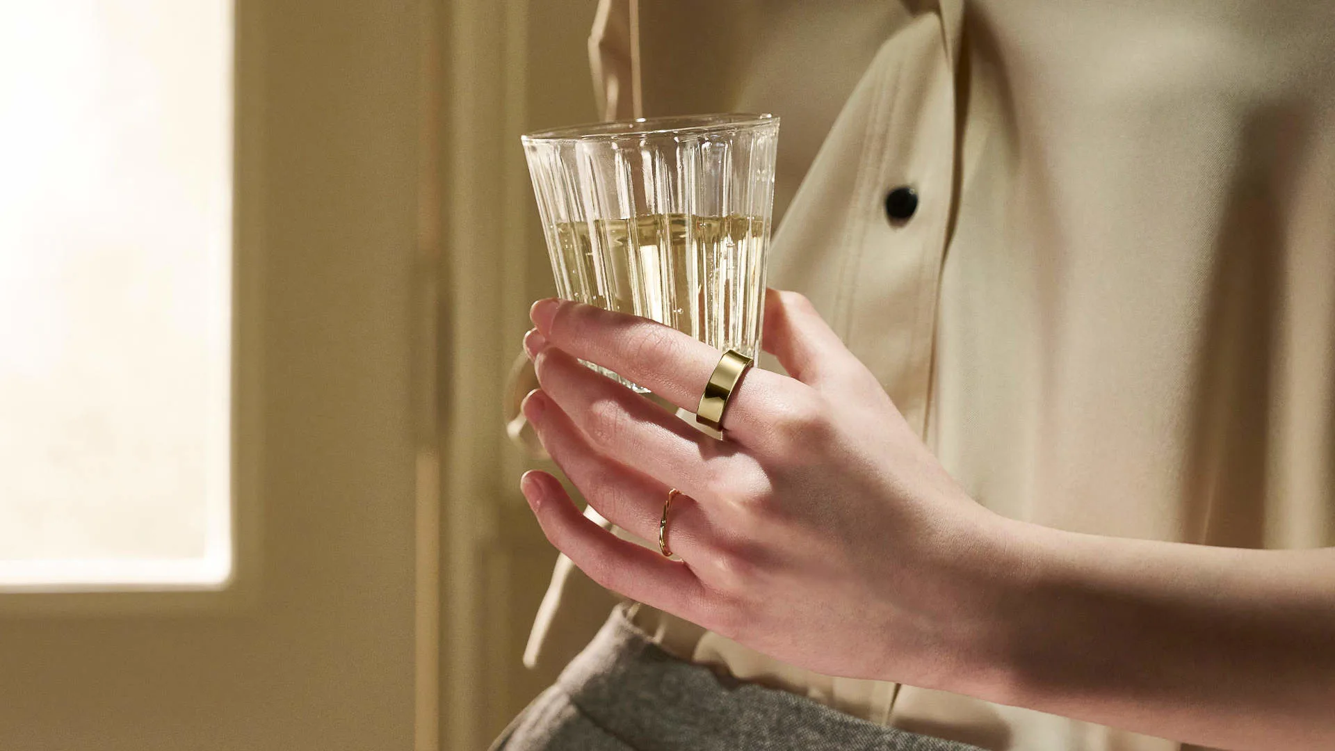 Een persoon houdt een glas met een heldere vloeistof vast, met gouden ringen om de vingers.