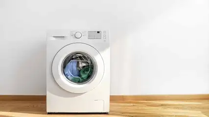 A white washing machine with a round door, sits against a white wall on a wooden floor. Clothes are visible inside the machine.