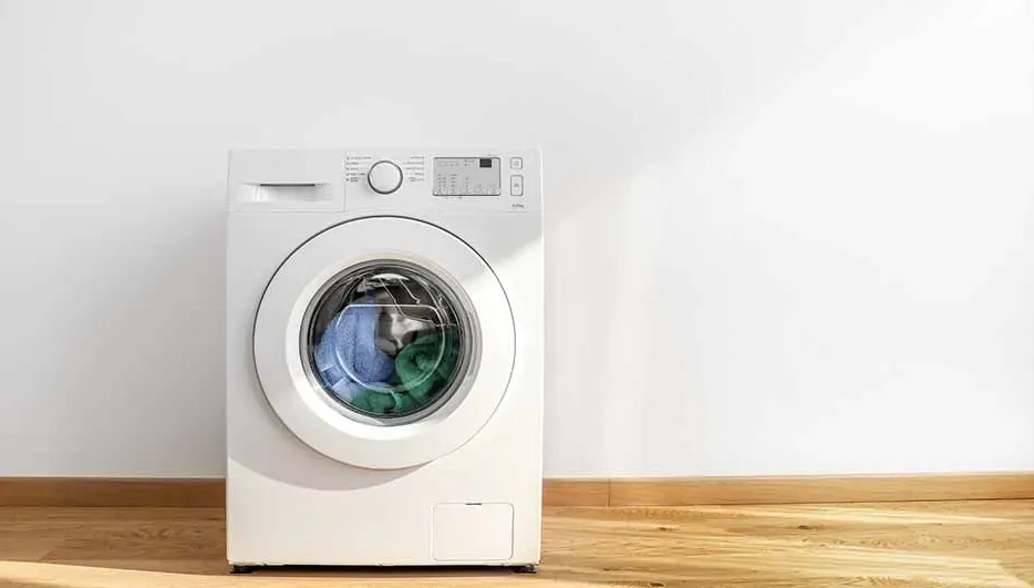 A white washing machine with a round door, sits against a white wall on a wooden floor. Clothes are visible inside the machine.
