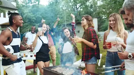 Een groep mensen staat buiten bij een barbecue met drankjes en een voetbal. Rook stijgt op van de grill.