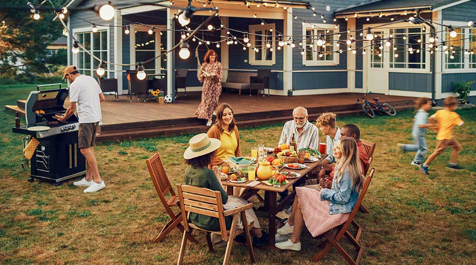Een familie geniet van een barbecue in de tuin, terwijl kinderen spelen en lichtslingers een warme, feestelijke sfeer geven.
