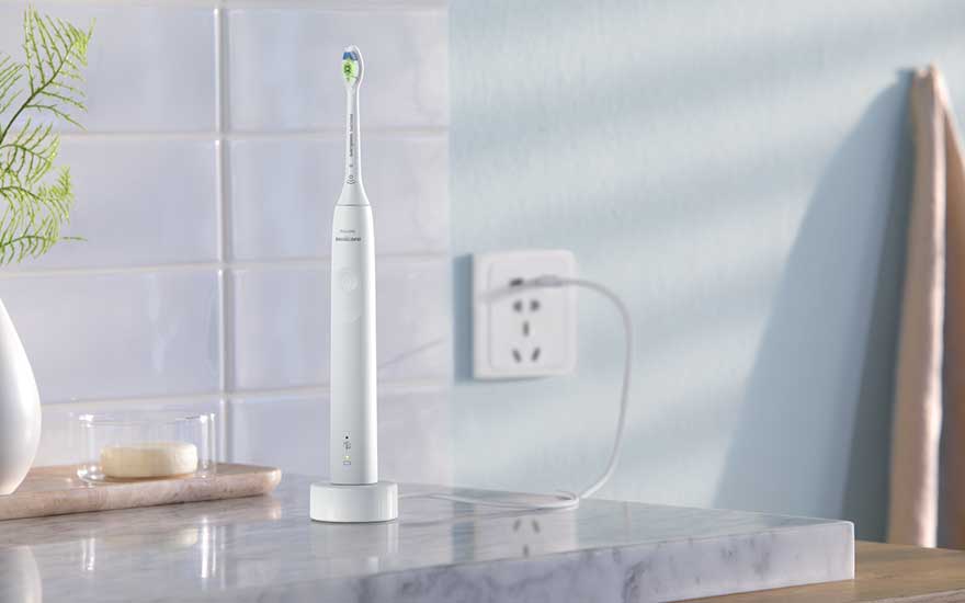 A white electric toothbrush stands on its charger atop a marble surface in a bright, tidy bathroom interior.