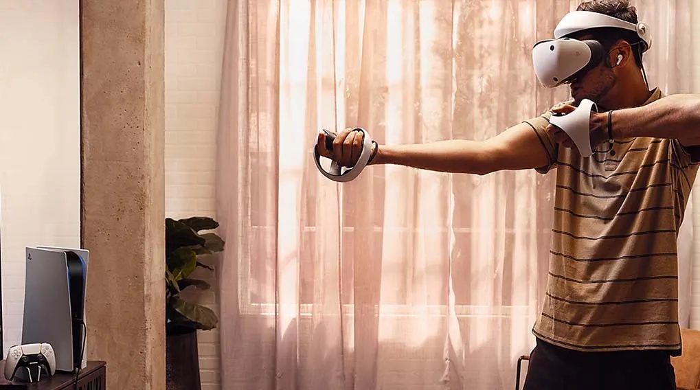 A person uses a VR headset and controllers in front of a curtained window. A game console and controller sit on a dark cabinet.