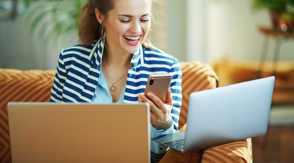 Cheerful woman in striped blazer sits on sofa with two laptops, smiling brightly as she reads a message on her smartphone.