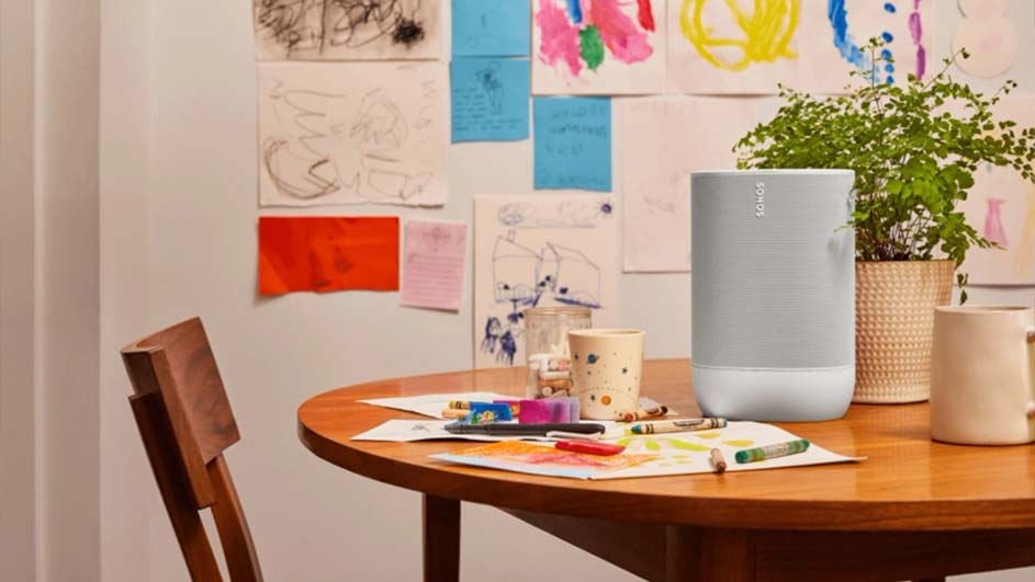 Ronde tafel met tekeningen, kleurpotloden, beker, plant, en een grijze speaker. Kindertekeningen aan de muur. Houten stoel links.