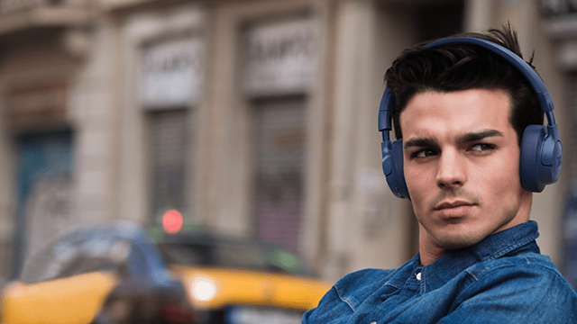 Jonge man met blauwe koptelefoon en spijkeroverhemd, kijkend naar rechts. Op de achtergrond gebouwen en een gele taxi.