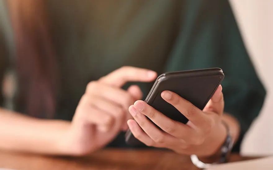 A person holds a black smartphone in both hands above a wooden table, one thumb resting while the other finger taps the bright unseen screen.