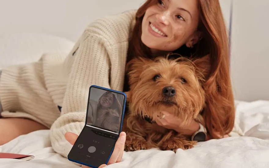Een vrouw ligt op bed met een kleine hond en gebruikt een blauwe klaptelefoon om een foto of selfie te maken in de slaapkamer.