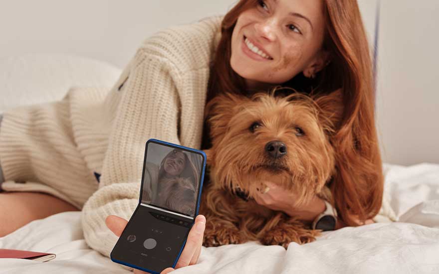 A woman lies on a bed with a small dog and uses a blue flip phone to take a photo or selfie in the bedroom.