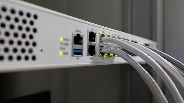 Een witte netwerkswitch met gele lampjes, zwarte poorten en grijze kabels is te zien tegen een grijze achtergrond.