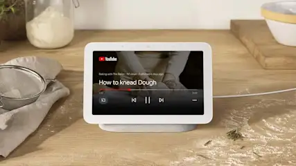 Een witte Google Nest Hub toont een YouTube-video over het kneden van deeg, met een zeef, bloem en een kom in de omgeving.