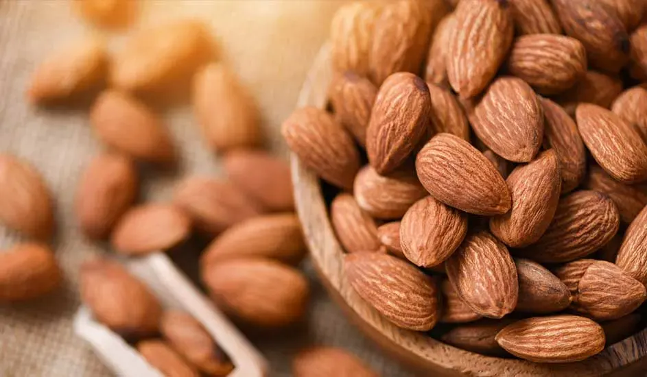 Een houten kom vol amandelen, sommige liggen ernaast op een tafel met een kleine witte lepel.