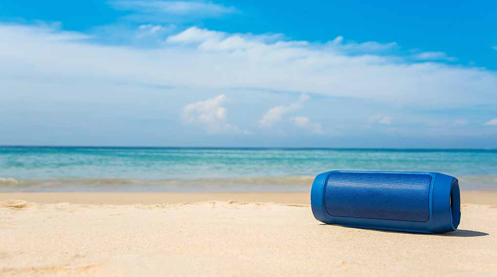 Een blauwe luidspreker ligt op een zandstrand. Achter de luidspreker is de blauwe zee en een blauwe lucht met witte wolken.