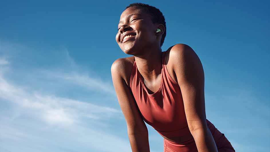 Lachende persoon in sportkleding met oortjes, tegen een blauwe lucht met wolken.