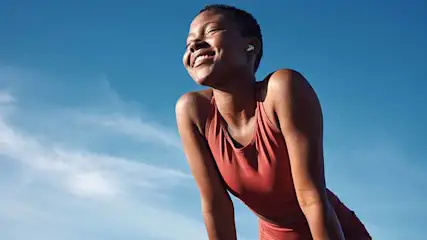 Lachende persoon in sportkleding met oortjes, tegen een blauwe lucht met wolken.