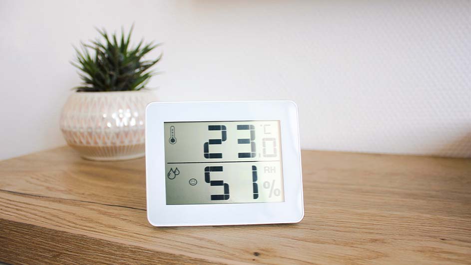 Een witte digitale thermometer toont 23 graden en 51% luchtvochtigheid op een houten tafel naast een plant.