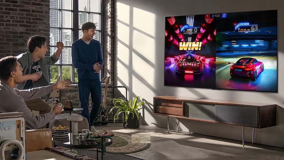 Drie mannen spelen een racegame op een groot scherm in een kamer met een bakstenen muur en een houten kast.