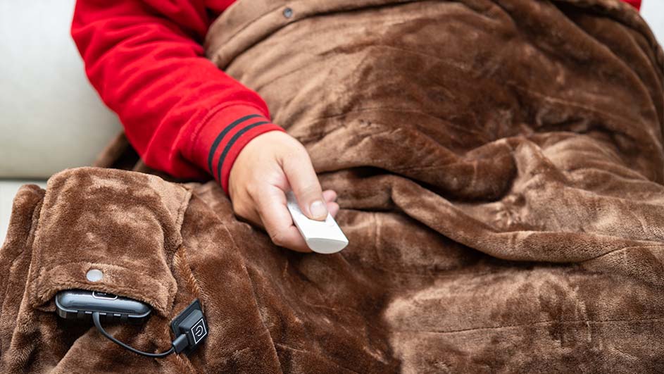 Een persoon onder een bruine deken houdt een witte afstandsbediening vast; een apparaat zit in een zak aan de deken.