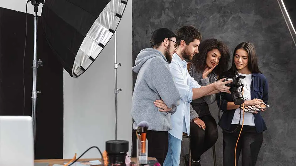 In een studio staan vier mensen rond een camera en een palet. Achter hen staat een lamp en een grijze achtergrond.