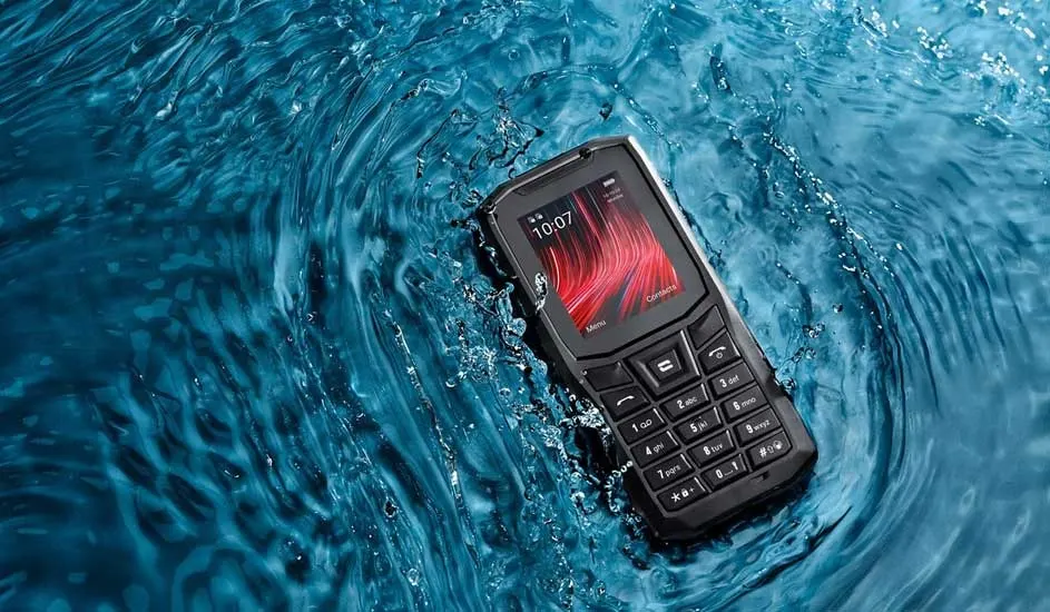A rugged black keypad phone floats in vivid blue water, ripples swirling around its small glowing screen showing red abstract lines.