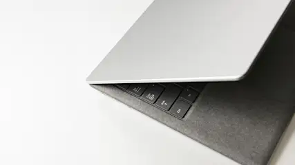 Een gedeeltelijk geopende, zilverkleurige laptop staat op een witte achtergrond, met zicht op het zwarte toetsenbord en grijze toetsenbordomgeving.