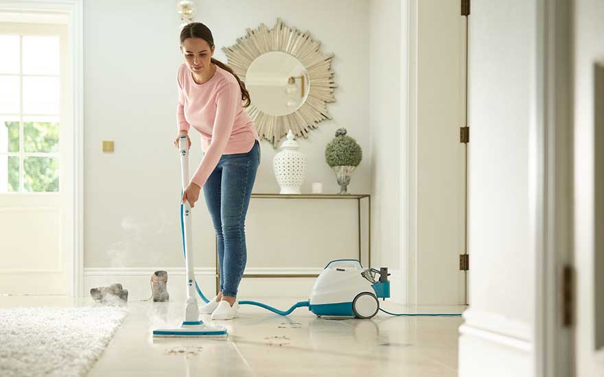 Jonge vrouw reinigt een tegelvloer met een stoomreiniger in een lichte hal; stoomwolken laten zien hoe hardnekkig vuil wordt verwijderd.