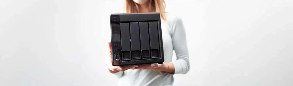 Een vrouw in een lichtgrijze trui houdt een zwarte NAS (Network Attached Storage) met vier schijfsloten voor zich vast. De achtergrond is egaal en lichtgrijs.