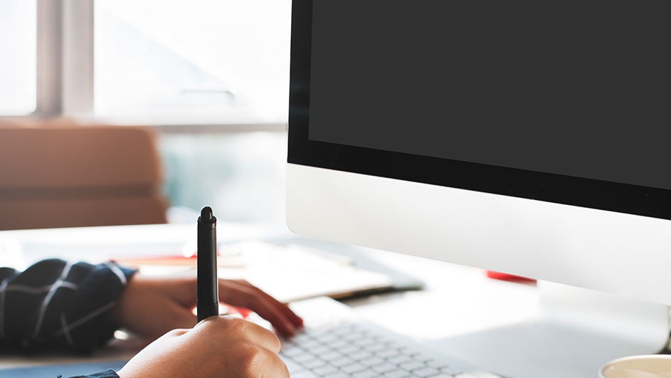 A person uses a black pen while typing on a white keyboard next to a computer with a black screen.