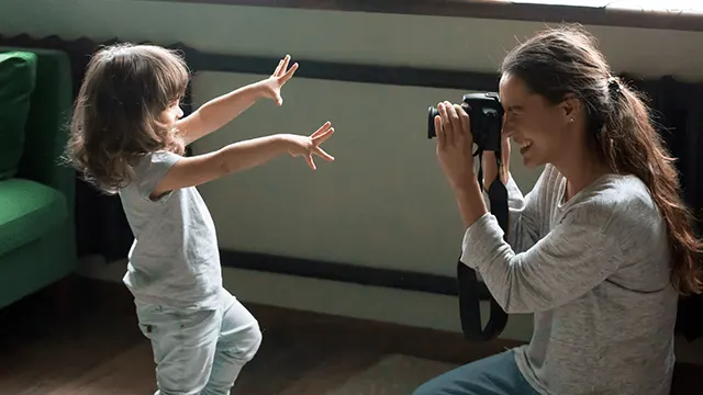 Een kind met opgestoken handen staat voor een vrouw met een camera in een kamer met een groene stoel.