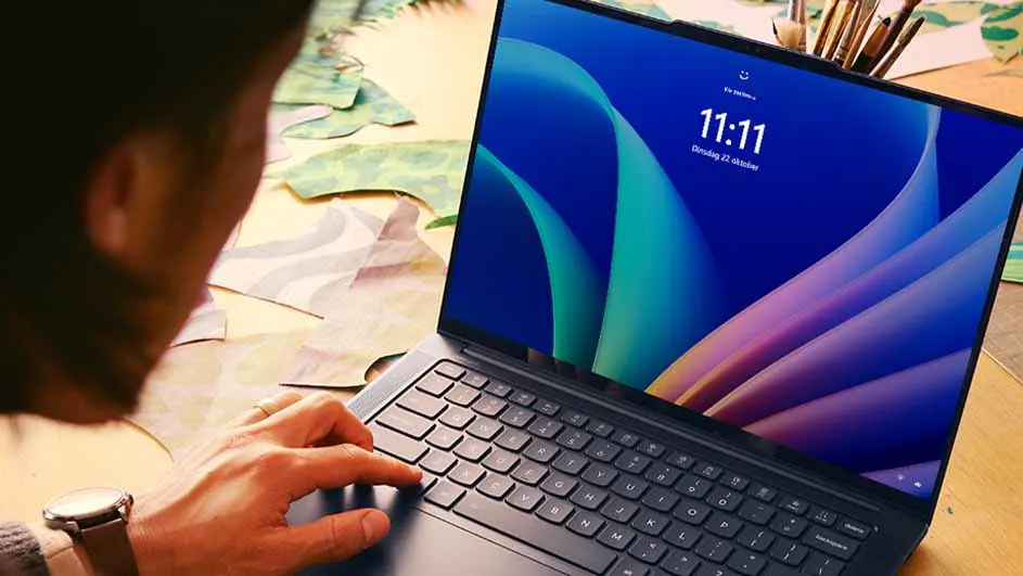 Een persoon typt op een laptop met een blauw scherm waarop de tijd 11:11 staat. Penselen staan rechts van het scherm.