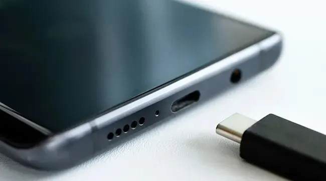 Een zwarte telefoon ligt naast een USB-C kabel op een witte ondergrond. De telefoon heeft een audio-aansluiting en speakergaatjes.