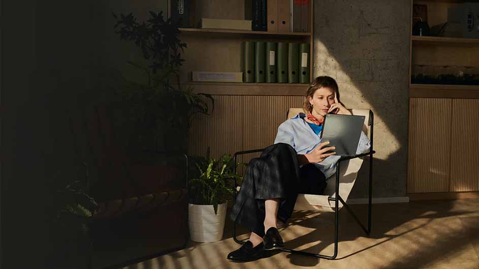 Vrouw zit in stoel met laptop, blauw hemd, donkere broek en schoenen. Achter haar een kast met ordners, plant en houten wand.