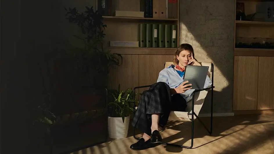 Vrouw zit in stoel met laptop, blauw hemd, donkere broek en schoenen. Achter haar een kast met ordners, plant en houten wand.